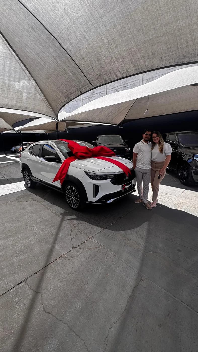 Casal ao lado de um carro branco com laço vermelho no capô, em concessionária