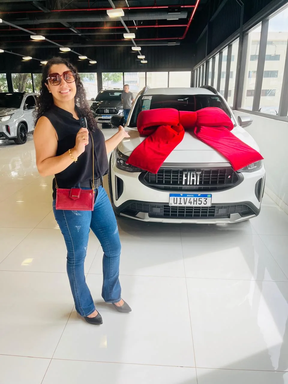 Mulher ao lado de um carro branco com laço vermelho no capô, em concessionária
