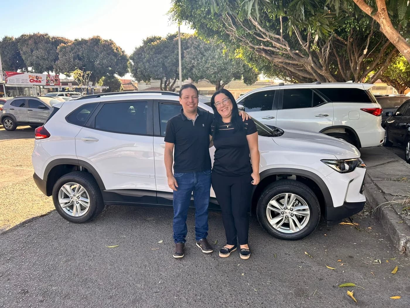 Casal ao lado de um SUV branco em estacionamento arborizado
